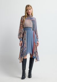 Mujer de pie con un vestido midi de cuello alto, estampado, con mangas largas y dobladillo asimétrico, combinado con botas negras hasta la rodilla.