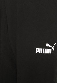 Calças de atletismo em algodão preto com um design ajustado. Apresenta um logo PUMA branco e um gráfico de puma a saltar no lado esquerdo. Textura suave.