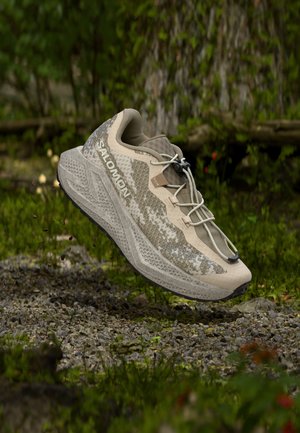 Μπεζ παπούτσι trail τρεξίματος Salomon με μαξιλαρωμένη σόλα αναρτημένο πάνω από βραχώδες, καλυμμένο με βρύα δασικό έδαφος.