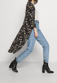 Kvinna som går i ljusblå jeans, svarta klackade ankelskor, vit topp och en lång svart kimono med blommönster.