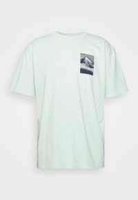 T-shirt à manches courtes vert menthe clair avec un petit motif de montagne en noir et blanc sur la zone de la poche poitrine gauche.