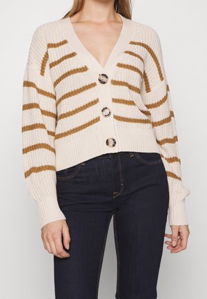 Frau trägt eine kurze cremefarbene Strickjacke mit braunen horizontalen Streifen und großen Knöpfen, kombiniert mit dunkelblauen, hoch taillierten Jeans.