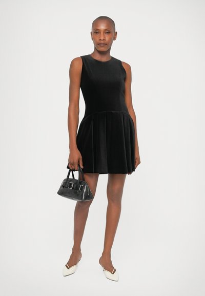 Zwart fluwelen jurk met een aansluitende lijfje en geplooide rok. Model houdt een kleine zwarte handtas vast. Witte puntige schoenen. Gladde textuur.