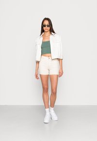 Modelo com um top verde cropped, uma jaqueta de ganga creme e calções creme, combinados com ténis brancos e óculos de sol verdes. Fundo simples.