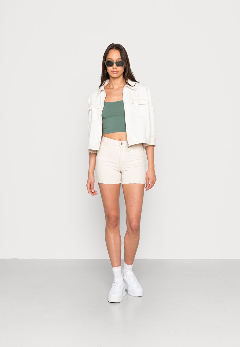 Modelo com um top verde cropped, uma jaqueta de ganga creme e calções creme, combinados com ténis brancos e óculos de sol verdes. Fundo simples.