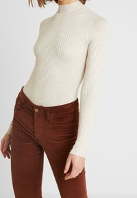 Femme portant un haut à manches longues côtelé beige à col roulé, associé à un pantalon en velours côtelé rouille taille haute, sur un fond uni.