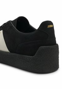 Baskets en daim noir avec une semelle plateforme épaisse et texturée, panneau latéral blanc, lacets et détail zigzag doré sur la languette du talon.