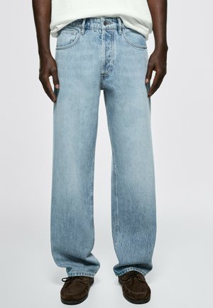 Personne portant un jean large bleu clair, une chemise blanche et des chaussures brunes, debout face à un fond uni.