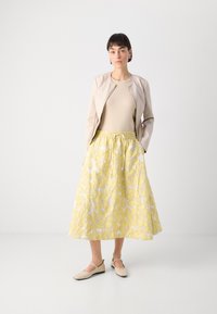 Veste en cuir beige sur un haut beige clair, assortie d'une longue jupe plissée jaune ornée de motifs floraux, et de chaussures beiges.