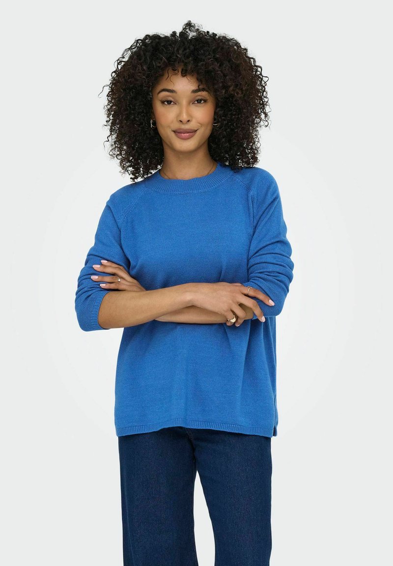 Blå stickad tröja med rundad halsringning och ribbade manschetter, i kombination med mörka jeans. Tyget verkar mjukt med en avslappnad passform.