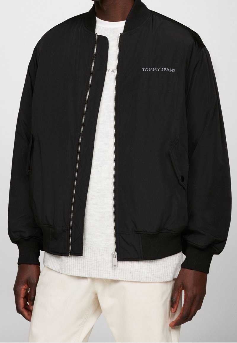 Homme portant une veste bomber noire zippée avec le logo « TOMMY JEANS » sur la poitrine, par-dessus un pull blanc et un pantalon crème.