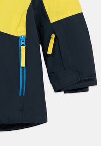 Námornícky modrá bunda s žltými akcentmi, obsahujúca veľkú žltú plochu, predný zips a bočné vrecká s jasnými zipsami. Štruktúrovaná tkanina.