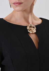 Schwarzes Kleid mit rundem Halsausschnitt, ausgestattet mit einer goldenen Rosenbrosche am Kragen. Glatte Textur, strukturiertes Design und dezente Details auf dem Stoff.