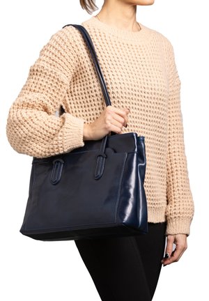 Marineblaue Leder-Tragetasche mit zwei Griffen, glatter Textur und kontrastierenden Nähten. Tragen von einer Person in einem cremefarbenen Strickpullover.