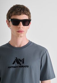 Crne naočale u obliku mačjih očiju s tamnim staklima, odmaraju se na licu modela. Model nosi sivi majicu s crnim logotipom.