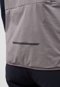 Leichte graue Jacke mit einer strukturierten Oberfläche, die einen dezenten schwarzen, reflektierenden Streifen an der Taille und einen elastischen Saum für einen sicheren Sitz aufweist.