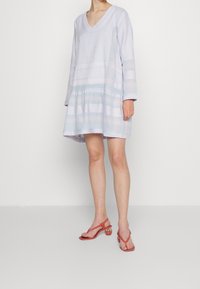 Jasnoniebieska sukienka do kolan z długimi rękawami, dekoltem w szpic, teksturowanymi wzorami i warstwowym dołem. Dobrana do różowych sandałów na paski.