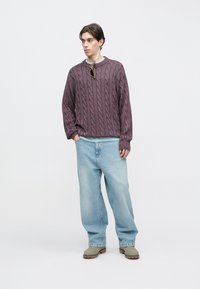 Μωβ πουλόβερ με πλεκτό σχέδιο και ανοιχτό μπλε γιακά, φορεμένο πάνω από ένα ανοιχτό μπλε πουκάμισο, σε συνδυασμό με άνετο ανοιχτό μπλε τζιν και γκρι μπότες.