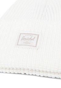 Bonnet en tricot blanc fabriqué à partir d'un tissu texturé avec un design à côtes, présentant une étiquette rectangulaire en beige clair avec des lettres foncées.