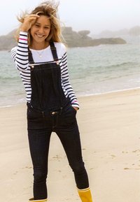 Denim overall met gouden accenten, gedragen over een zwart-witte gestreepte longsleeve shirt, gecombineerd met gele regenlaarzen op een zandstrand.