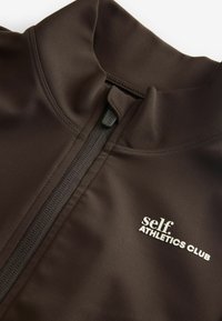 Must musta jakk, millel on kõrge krae, täispikk tõmblukk ja valge trükitud logo, mis ütleb "self. ATHLETICS CLUB". Sile, veniv kangas.