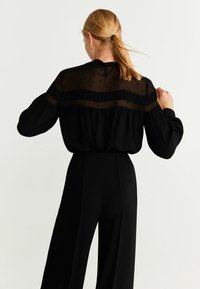 Blusa nera in tessuto trasparente con motivo a pois, maniche lunghe a sbuffo e vestibilità comoda, abbinata a pantaloni neri a vita alta.