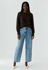 Pull en maille marron, jean large bleu clair avec détails en patch, et escarpins blancs. Le mannequin a de longs cheveux tressés.