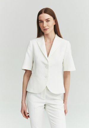 Młoda kobieta o długich brązowych włosach, ubrana w biały żakiet z krótkim rękawem i pasujące białe spodnie na jasnym tle.