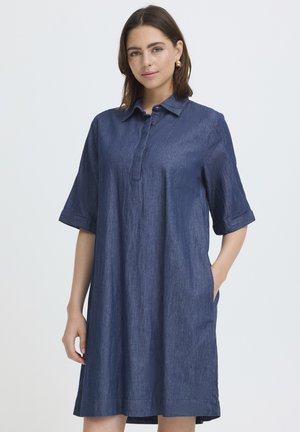 Femme aux cheveux bruns mi-longs portant une robe chemise en denim bleu foncé à manches courtes et amples, debout avec une main dans la poche.