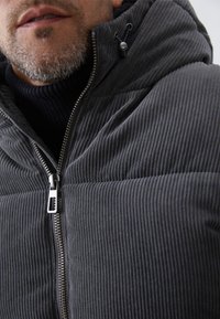 Grau gestreifte Cordjacke mit Reißverschluss und Kapuze, die eine strukturierte Oberfläche und einen hohen Kragen über einem dunklen Pullover aufweist.