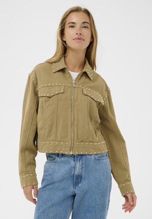 Femme portant une veste courte de couleur beige avec des clous argentés, fermeture éclair à l'avant, et un jean en denim bleu sur un fond blanc.