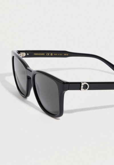 Occhiali da sole neri a forma di occhio di gatto con lenti grigie, finitura opaca, dettagli in metallo argento e logo distintivo sulla tempia.