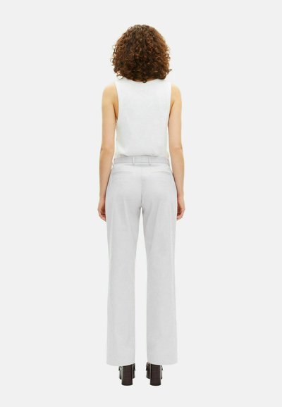 Vrouw met krullend haar, in een witte mouwloze top, hooggepassen witte broek en zwarte hoge hakken, staand met de rug naar de camera.