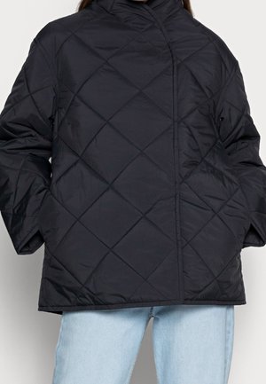 Veste matelassée noire avec couture en losanges portée sur un jean bleu clair, mains dans les poches, vue de face recadrée au niveau du cou et à mi-cuisse.