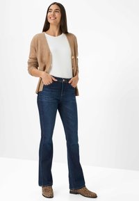 Cardigan beige con bottoni, top bianco, jeans a zampa d'elefante blu scuro e stivaletti alla caviglia con stampa leopardata marrone, in piedi con le mani in tasca.