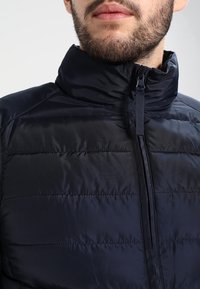 Dunkelblauer Steppmantel mit hohem Kragen, horizontalem Steppmuster und Frontreißverschluss. Glattes, leichtes Material mit glänzendem Finish.