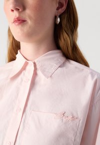 Camicetta button-up rosa chiaro con colletto a punta, dotata di tasca frontale e logo ricamato sottile in filo abbinato. Tessuto liscio.