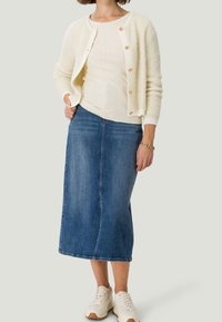 Weißer, flauschiger Cardigan über einem gerippten cremfarbenen Oberteil, kombiniert mit einem dunkelblauen Denim-Midirock und weißen Sneakers mit beigen Akzenten.
