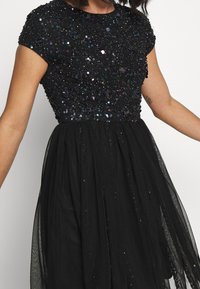 Svart klänning med korta ärmar, beaded bodice dekorerad med iriserande paljetter, flödande genomskinlig kjol och strukturerade tygdetaljer.