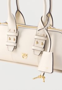 Cremefarvet lædertaske med guldbelagte metaldele, dobbelte håndtag og justerbare remme. Har et logoemblem og en hængende mærkat med nøgler.