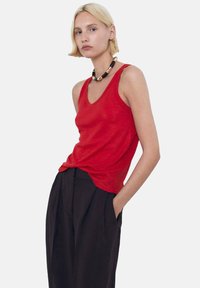 Red sleeveless tank top in a lightweight fabric, paired with high-waisted black trousers. Notable necklace features black and gold details.