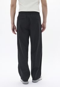 Osoba stoi w luźnych czarnych spodniach, białej bezrękawnikowej koszulce i białych trampkach, pokazana z tyłu na jednolitym tle.