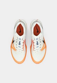 Fehér és narancssárga sportcipők texturált hálós felsőrésszel, fekete Nike logóval, narancssárga díszítéssel és fűzőkkel, amelyek a nyelvön keresztül húzódnak.