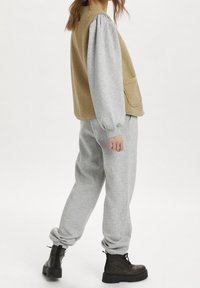 Hnědá sherpa vesta přes šedou mikinu a tepláky. Outfit má měkkou texturu s volným střihem a manžetami na nohavicích. Černé kotníkové boty.