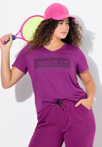 Livlig lilla træningsoutfit bestående af en V-hals T-shirt med sort tekst og matchende joggingbukser, suppleret med en pink kasket og en tennisracket.
