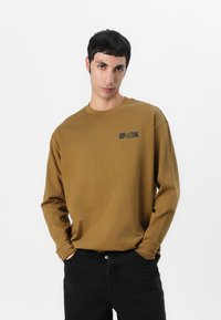 Brown long-sleeve t-shirt with a crew neck and black logo on the chest, paired with black pants; features a relaxed fit and soft texture.