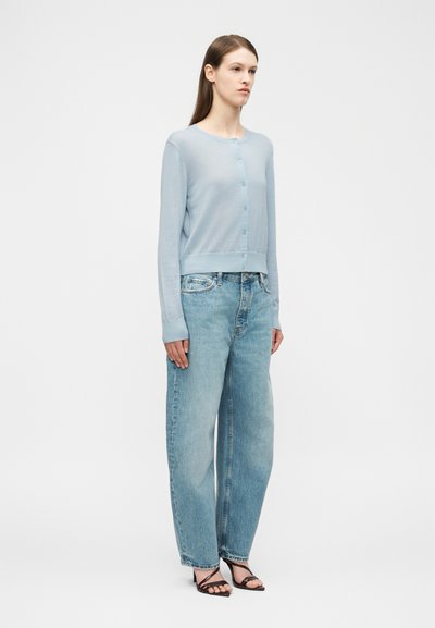 Vrouw staand, met een lichtblauwe gebuttonde cardigan, losse lichtblauwe jeans en zwarte sandaaltjes met hakken, voor een effen witte achtergrond.