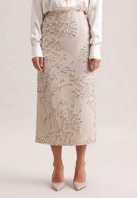 Gonna midi beige con motivi floreali ricamati in argento, abbinata a una blusa in satin bianca e décolleté beige a punta.
