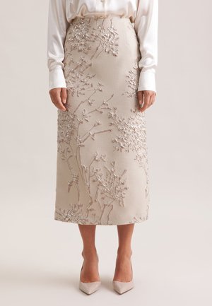Gonna midi beige con motivi floreali ricamati in argento, abbinata a una blusa in satin bianca e décolleté beige a punta.