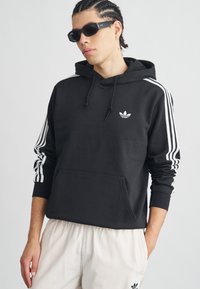 Juodas bliuzonas su gobtuvu, ant krūtinės baltas Adidas logotipas ir baltos trijų juostelių dėmės ant rankovių. Minkšta medžiaga, kišenė priekyje kaip kanguros kišenė.